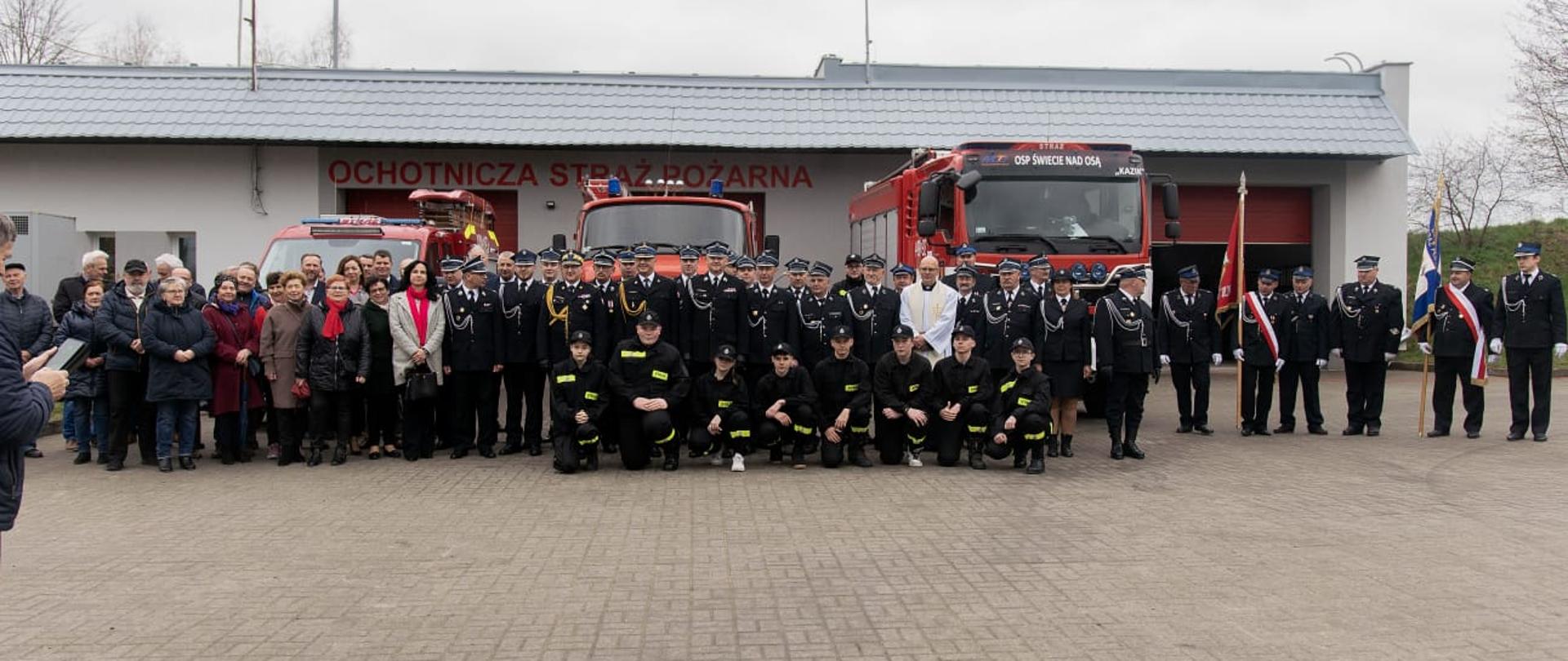Ustawieni do zdjęcia grupowego strażacy i zgromadzeni ludzi na tle pojazdów pożarniczych i jednostki OSP