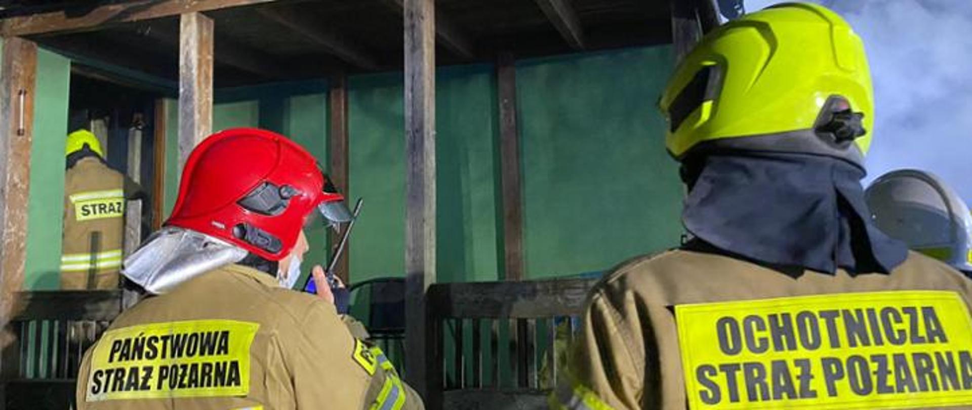 Strażak PSP po lewo i strażak OSP po prawo, widok z tyłu, przed nimi drewniany budynek i widoczne zadymienie