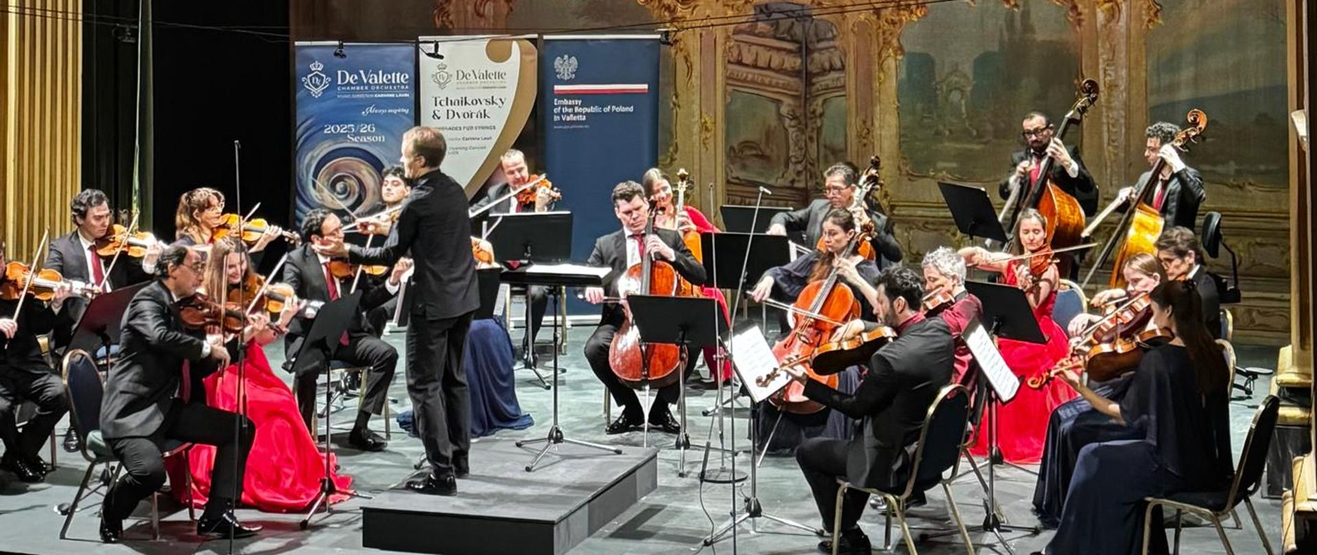A chamber orchestra on stage conducted by Christoph Koncz. Women are wearing red dresses, men in black suits, white shirts, and pink ties. In the background, roll-ups of the Polish Embassy in Valletta and the De Vallette Chamber Orchestra are visible. The photo is taken from a close distance.