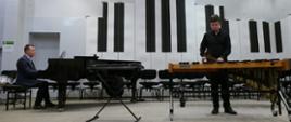 Chłopak stojąc na estradzie auli PSM gra na ksylofonie. Po lewej stronie mężczyzna gra na fortepianie.
