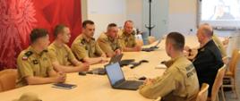 Wnętrze Sali konferencyjnej. Przy owalnym stole, na którym leżą przenośne komputery, siedzą mężczyźni, strażacy w umundurowaniu służbowym. Za nimi biały ekran z obrazem transmisji on-line (zdjęcie mężczyzny w czarnej koszulce). 