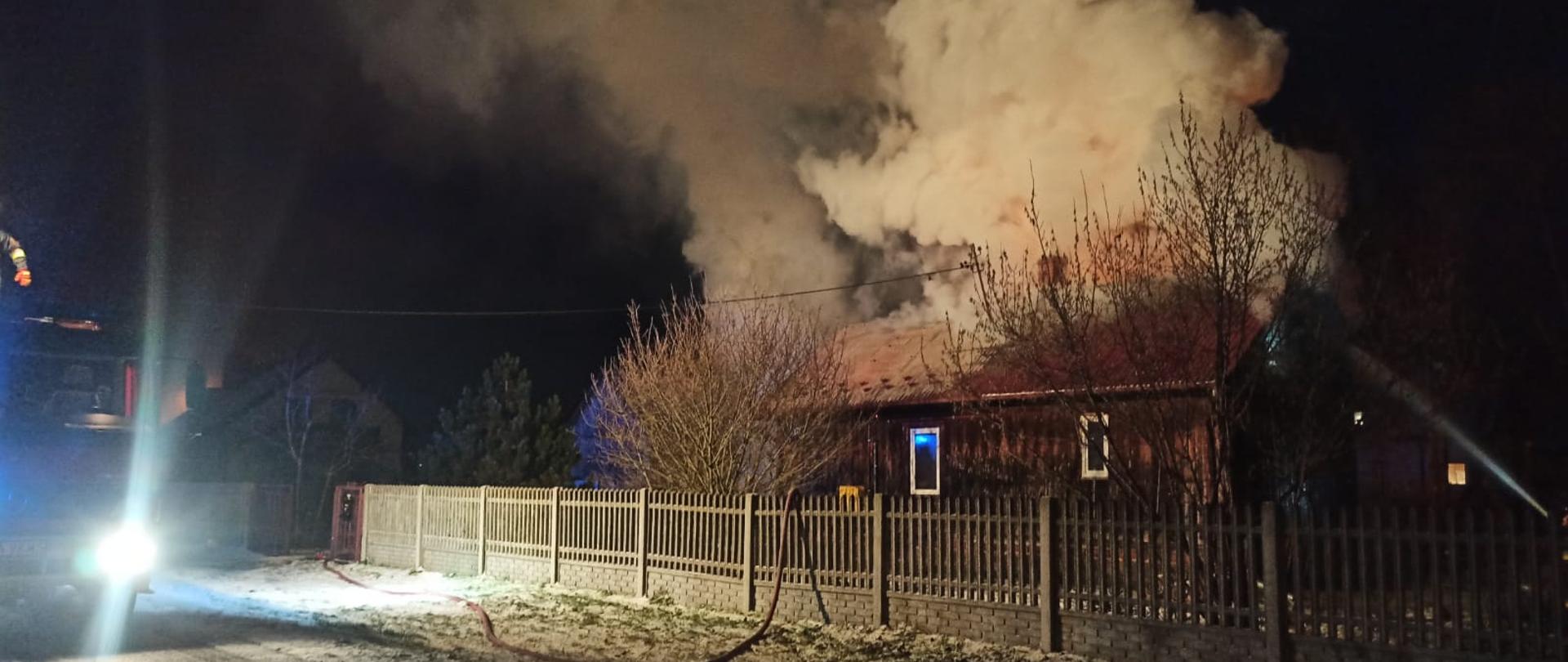 Na zdjęciu widać dom drewniany objęty pożarem i samochód pożarniczy stojący na drodze.