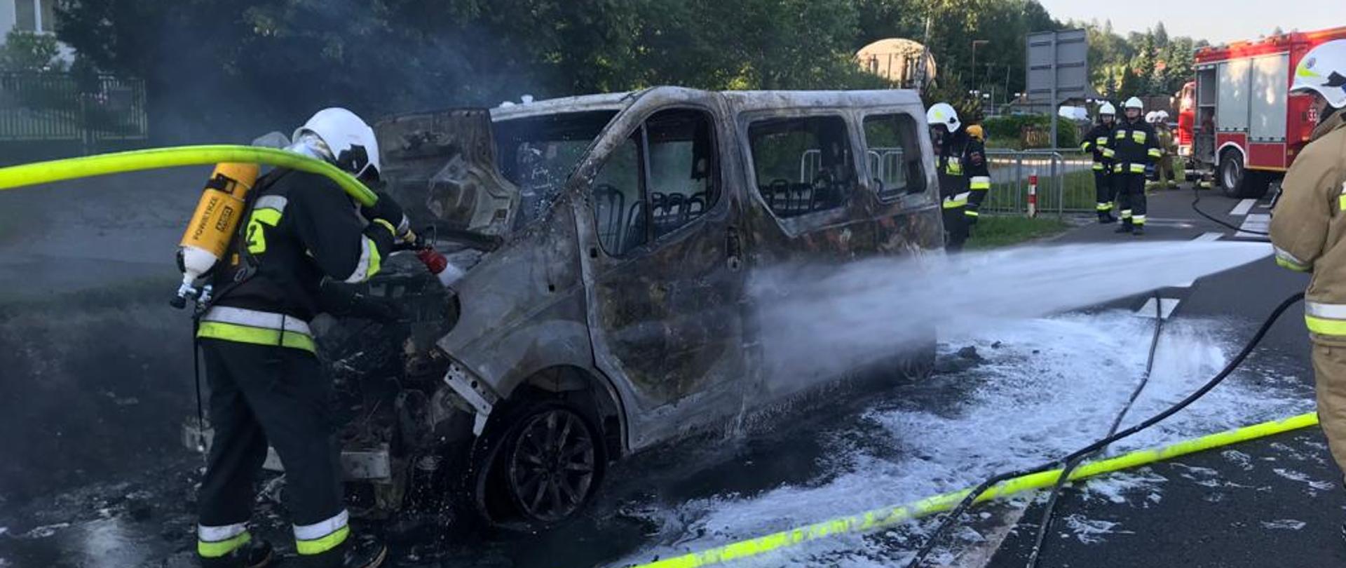 Na zdjęciu widzimy spalony samochód przy którym strażacy prowadzą działania gaśnicze. Zdarzenie ma miejsce na poboczu drogi krajowej nr 7. Z prawej strony widoczne są samochody strażackie i pozostali strażacy. Tło zdjęcia obejmuje zabudowania miejscowości oraz krzewy i drzewa. 