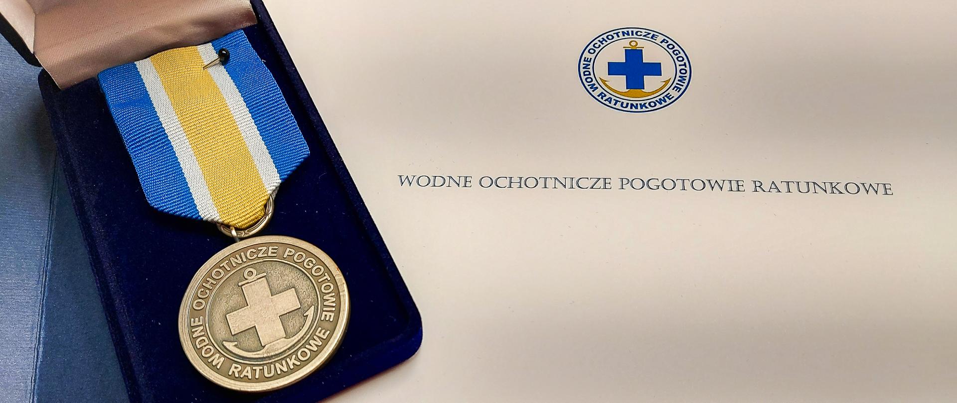 Zdjęcie przedstawia srebrny medal za zasługi dla Wodnego Ochotniczego Pogotowia Ratunkowego. Medal umieszczony jest na teczce z napisem i Logotypem WOPR.