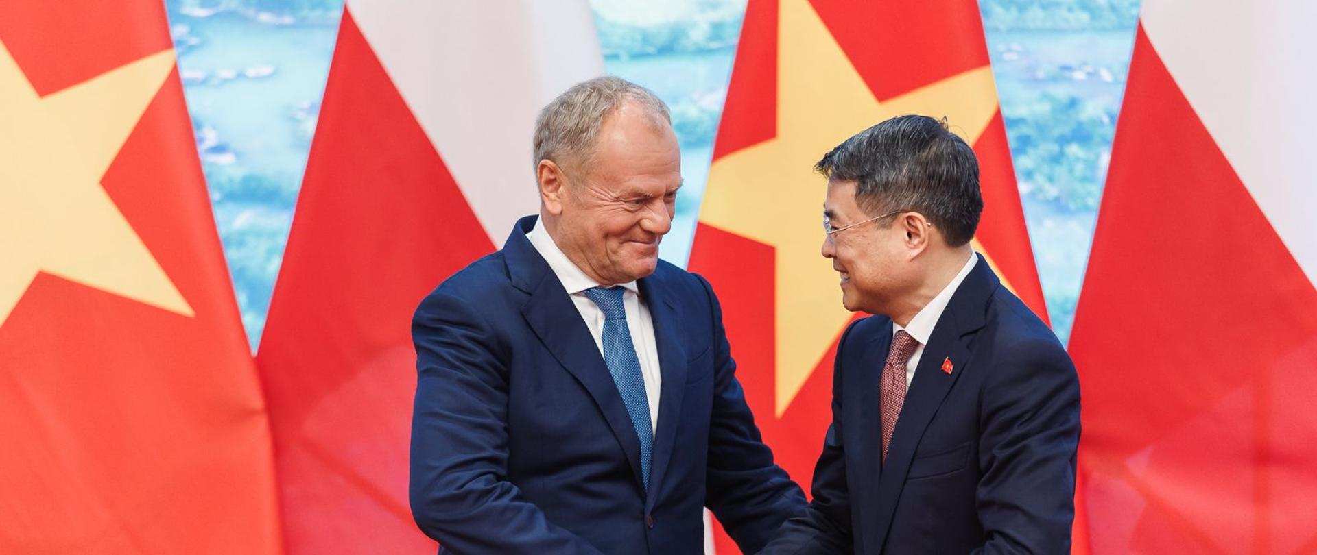 Premier Polski Donald Tusk i premier Wietnamu Lê Minh Hưng witają się uściskiem dłoni podczas oficjalnego spotkania. W tle widoczne są flagi Polski i Wietnamu.