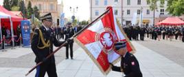 Poczet sztandarowy Państwowej Straży Pożarnej podczas ceremonii. Jeden ze strażaków klęczy przed sztandarem, a pozostali funkcjonariusze stoją na czerwonym dywanie.