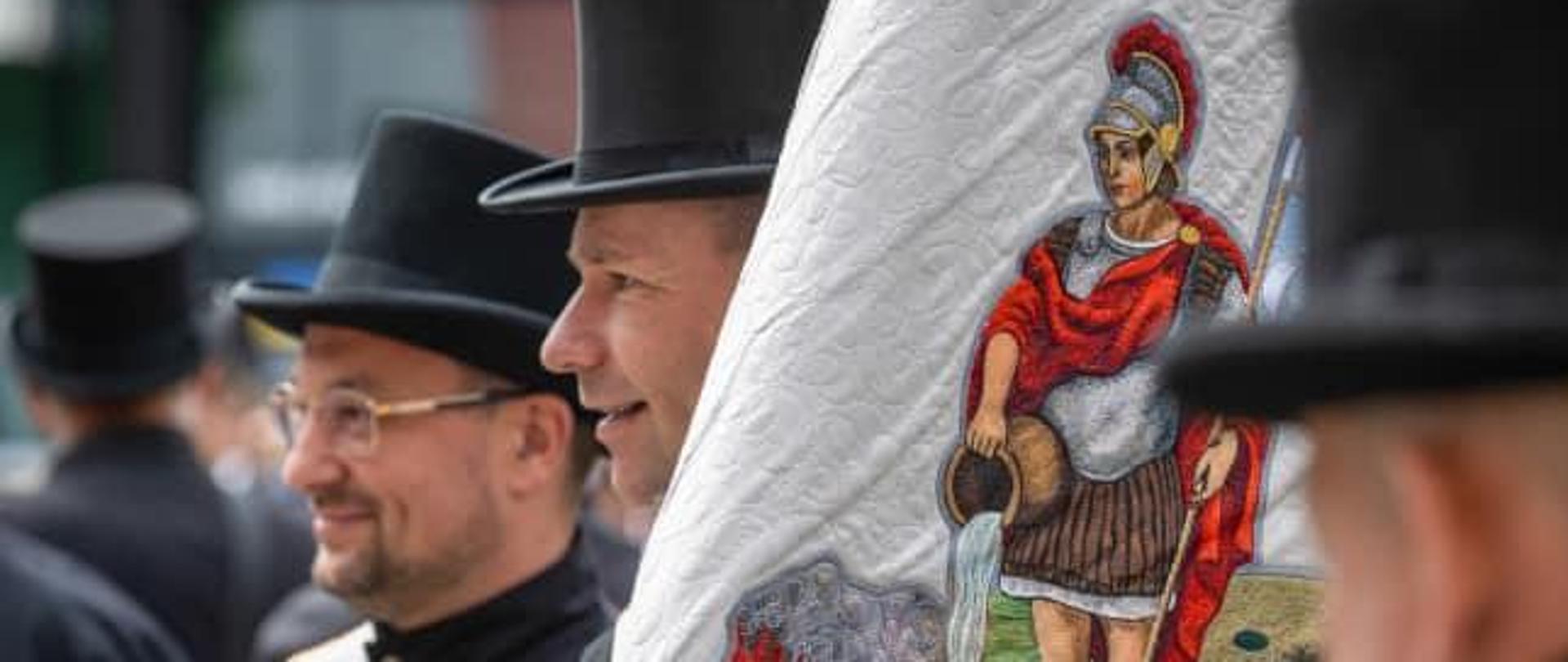 
Widok na fragment sztandaru z wizerunkiem świętego Floriana. Obok widać kilku kominiarzy w mundurach galowych i w cylindrach.
