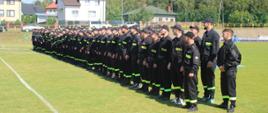 
Oto alternatywne opisy zdjęć dla osób niewidomych, bazujące na informacjach o zawodach sportowo-pożarniczych OSP w gminie Wąsewo:
Zdjęcie 1:
To zdjęcie przedstawia długi szereg strażaków ochotników, ustawionych równo na zielonym boisku sportowym. Wszyscy ubrani są w ciemne kombinezony z jaskrawymi, prawdopodobnie zielonymi lub żółtymi, paskami odblaskowymi, oraz w ciemne czapki z daszkiem. Stoją w lekkim skosie, patrząc w jednym kierunku. W tle widać domy mieszkalne i las, a niebo jest bezchmurne. Scena sugeruje uroczyste otwarcie lub apel.