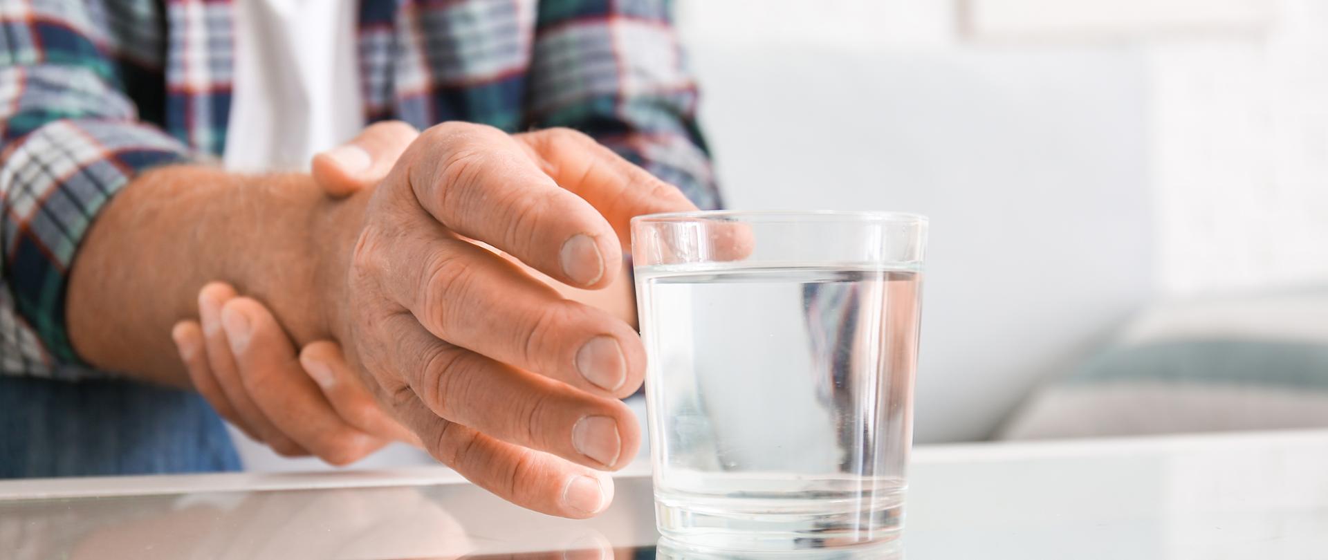 Senior man with Parkinson syndrome taking glass of water from table, closeup