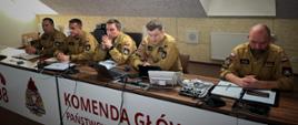 Grupa strażaków PSP w mundurach koloru żółtego siedzi przy stole w sali operacyjnej Komendy Głównej PSP. Na stole znajdują się laptopy, mikrofony oraz dokumenty, a w tle widać logo PSP i numer alarmowy 998.