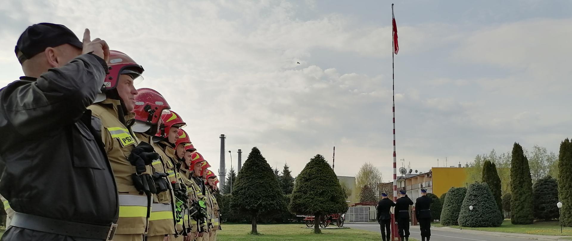 Na zdjęciu widać strażaków ustawionych w dwuszeregu, którzy oddają honor podczas uroczystego podniesienia flagi. Na drugim planie stoi poczet flagowy