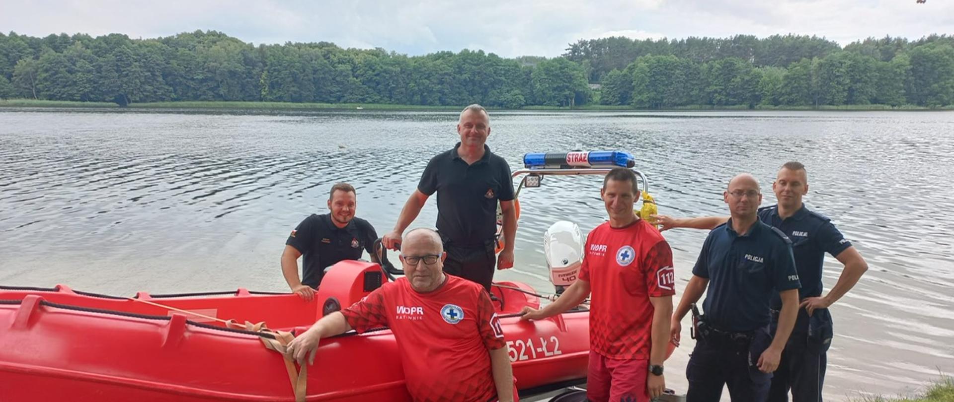 Zdjęcie wykonano w ciągu dnia nad akwenem wodnym, jeziorem, na pierwszym planie łódź ratunkowa motorowa, przy niej dwóch strażaków państwowej straży pożarnej, dwóch funkcjonariuszy policji i dwóch ratowników wodnego ochotniczego pogotowia ratunkowego pozuje do zdjęcia. 