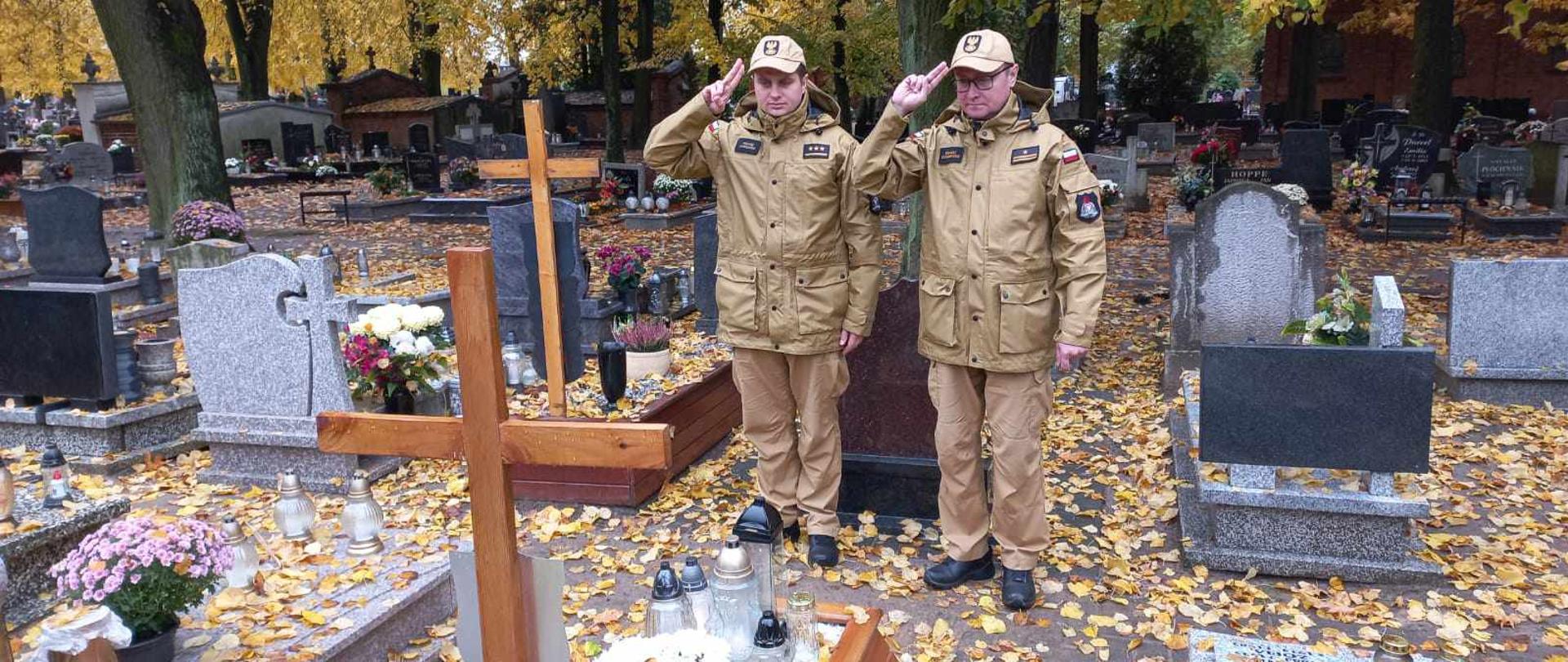 Zdjęcie przedstawia dwóch strażaków w mundurach służbowych salutujących nad grobem
