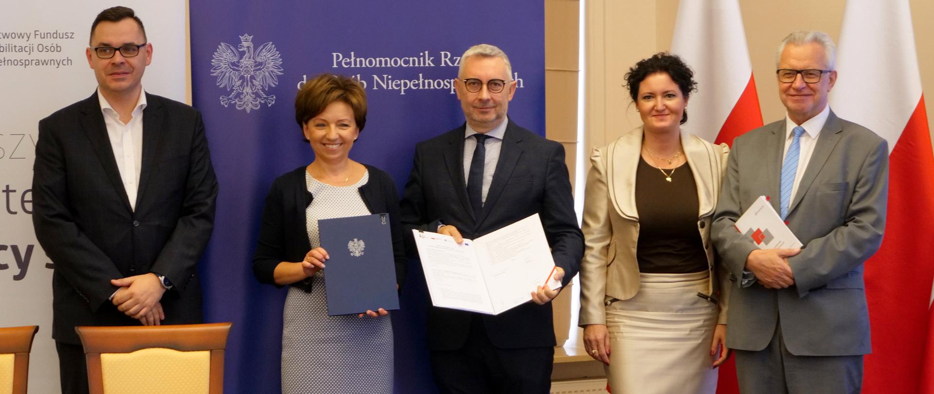 W sali za stołem stoi pięć osób. Dwie z nich prezentują dokumenty. Za nimi stoją flagi Polski i baner PFRON.