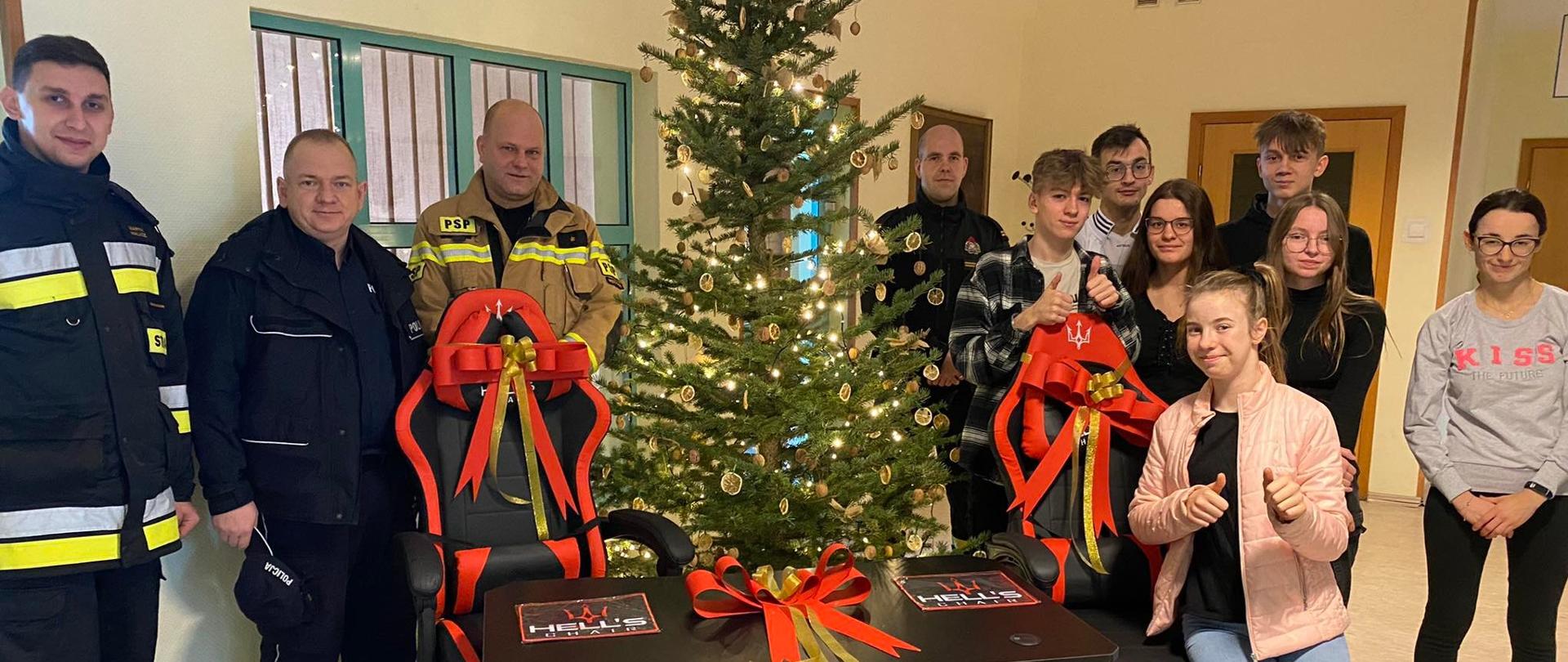 Na zdjęciu strażacy, policjant i wychowankowie placówki w Chrzypsku stoją przy choinki, przed która znajdują się dwa biurka i dwa fotele gaminkowe - prezenty dla placówki w Chrzypsku.