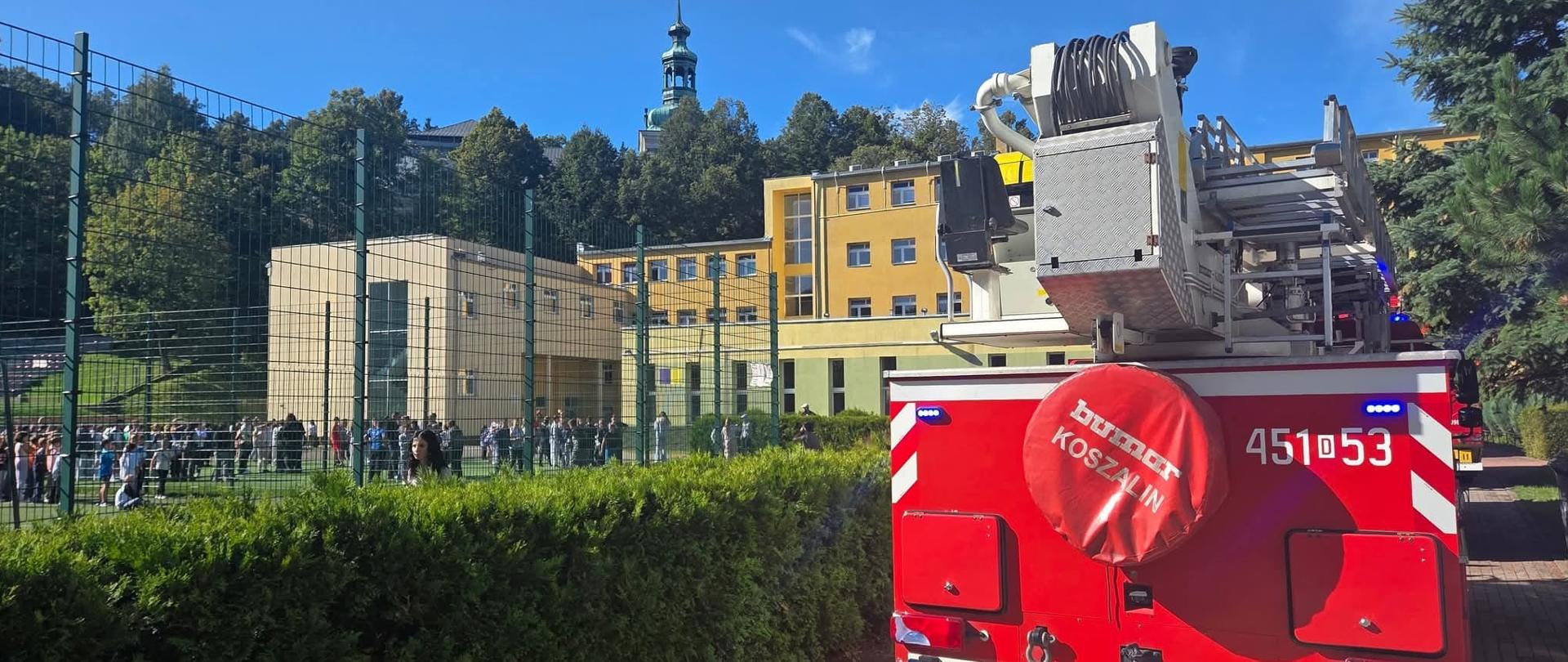 W tle budynek szkoły podstawowej na planie głównym pojazd straży pożarnej. Po lewej stronie dostrzegamy plac, na którym znajdują się uczniowie ewakuowani z zabudowań.