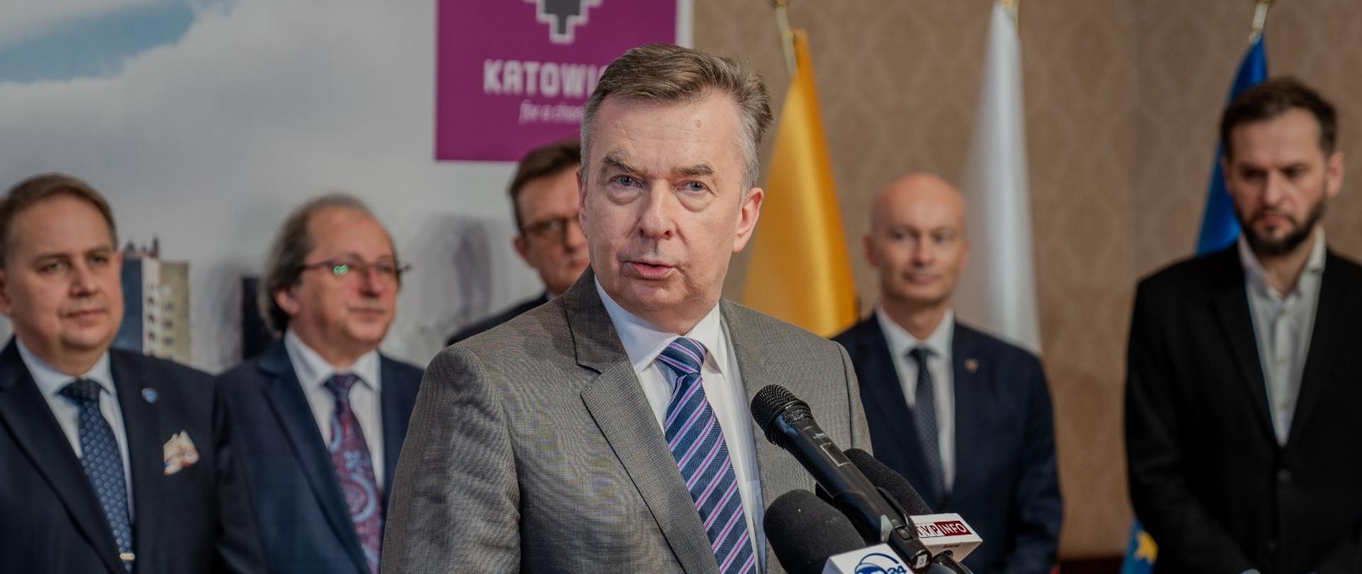 Minister Wieczorek stoi i mówi do kilku mikrofonów, za nim kilku mężczyzn w garniturach.