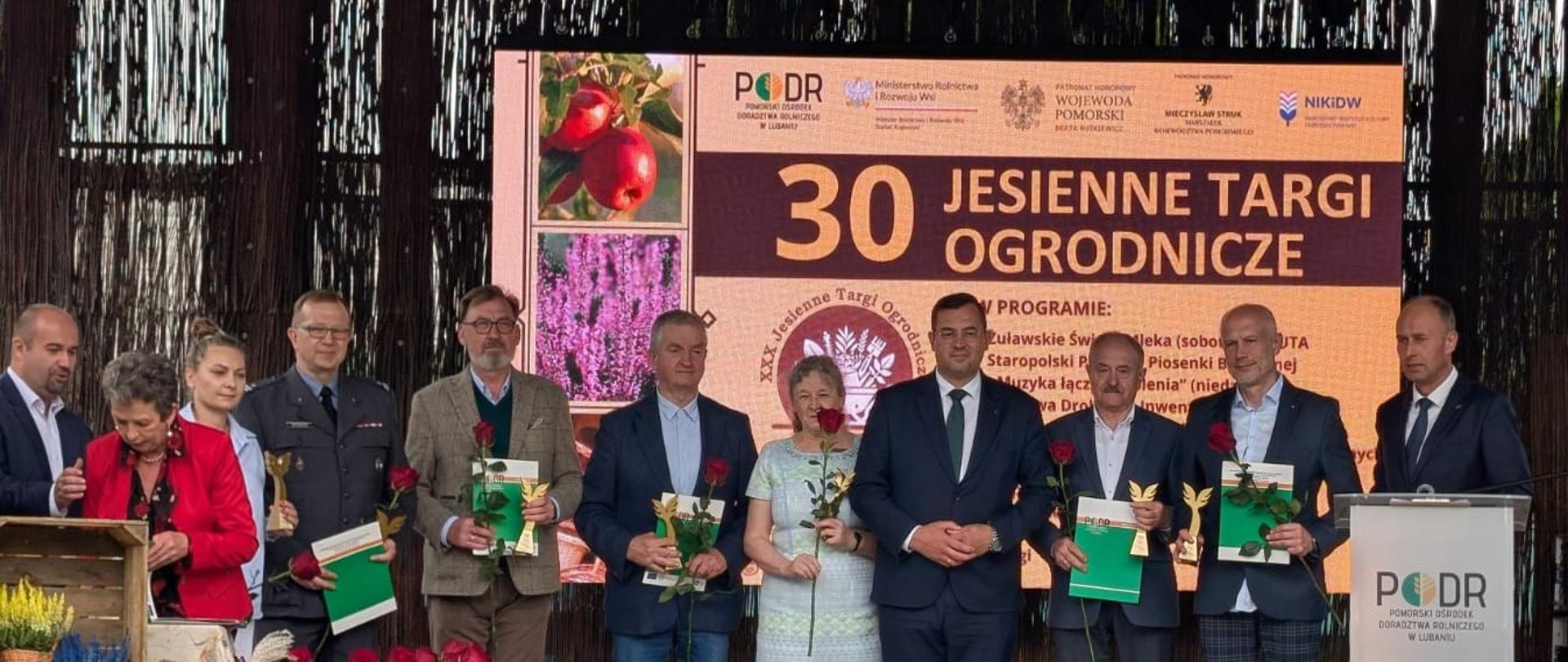 Na scenie grupa osób – laureaci i organizatorzy wydarzenia – trzymający dyplomy, statuetki oraz róże. W tle widoczna jest duża tablica z nazwą wydarzenia oraz logotypami instytucji wspierających targi. Atmosfera zdjęcia jest uroczysta i oficjalna.