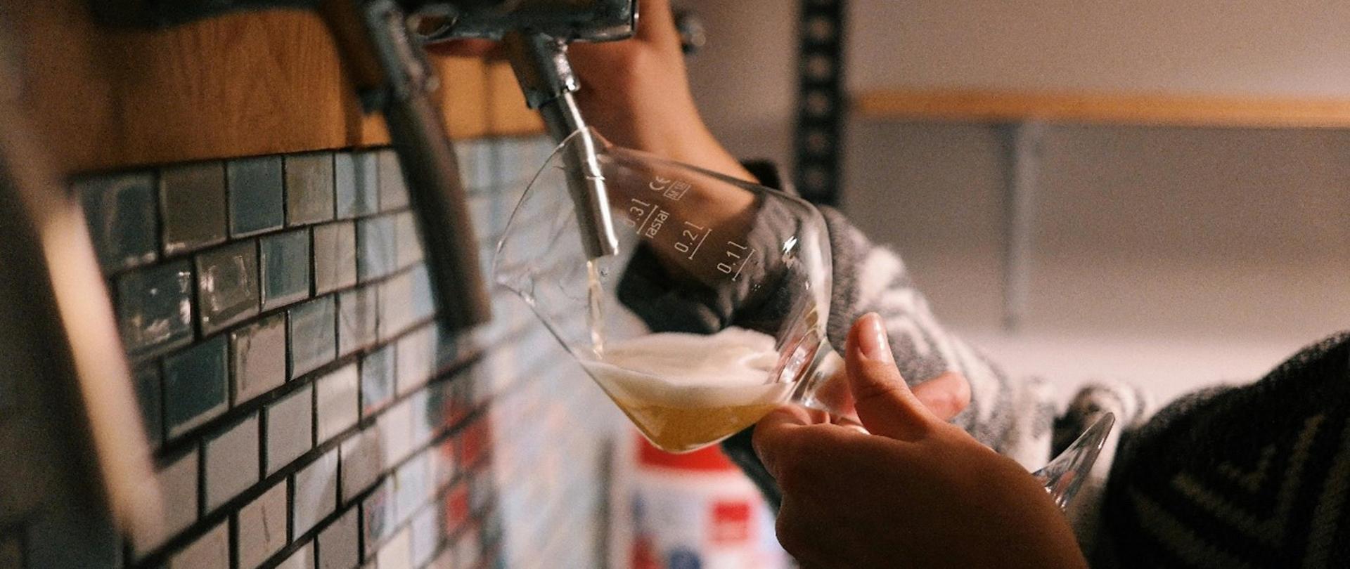 Zdjęcie przedstawia dłoń która nalewa piwo do szklanki.