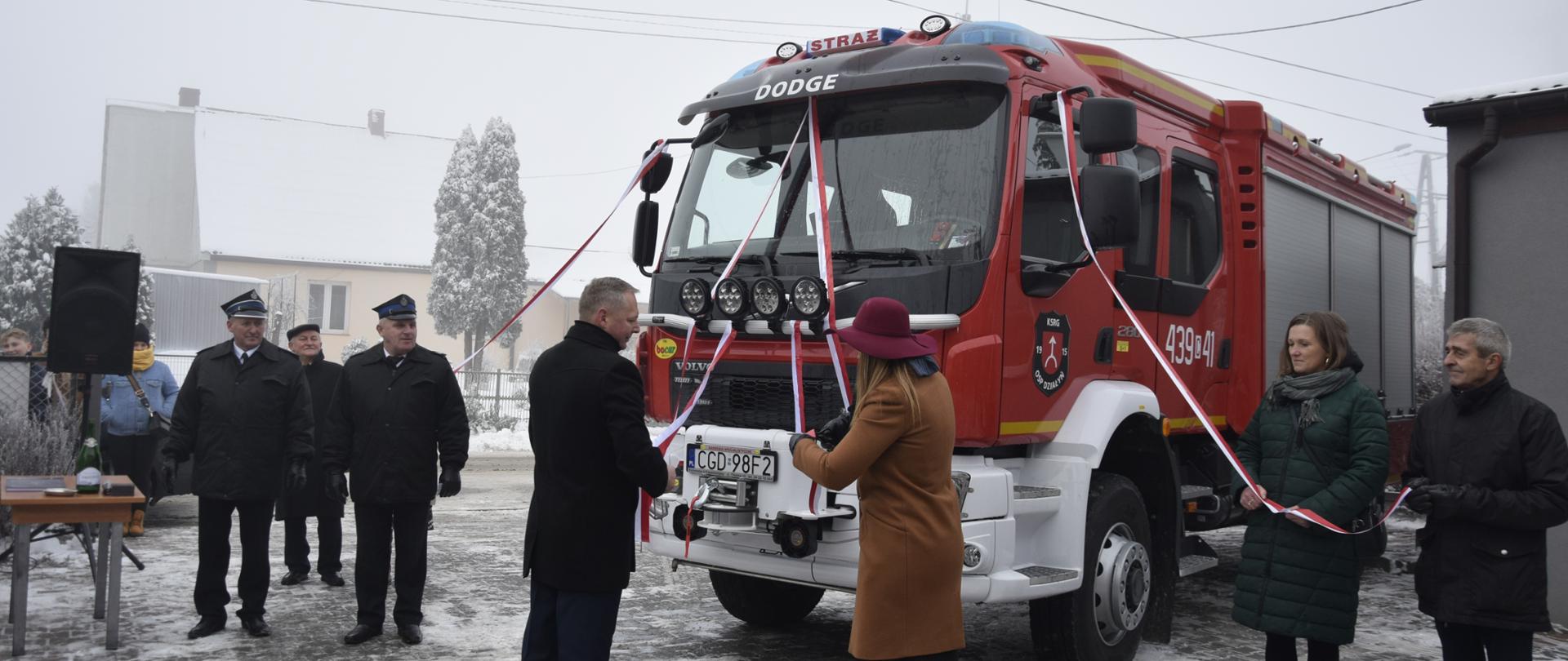 Przy nowym wozie strażackim stoją kobiety i mężczyźni i trzymają w dłoniach szarfy ozdobne w kolorze biało-czerwonym