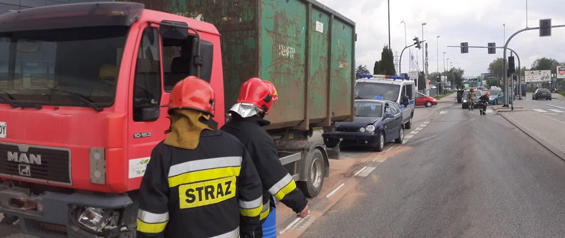 Samochody uczestniczące w zdarzeniu stoją na poboczu. samochód ciężarowy, osobowy i radiowóz Policji. Strażacy zasypują rozlane płyny sorbentem.