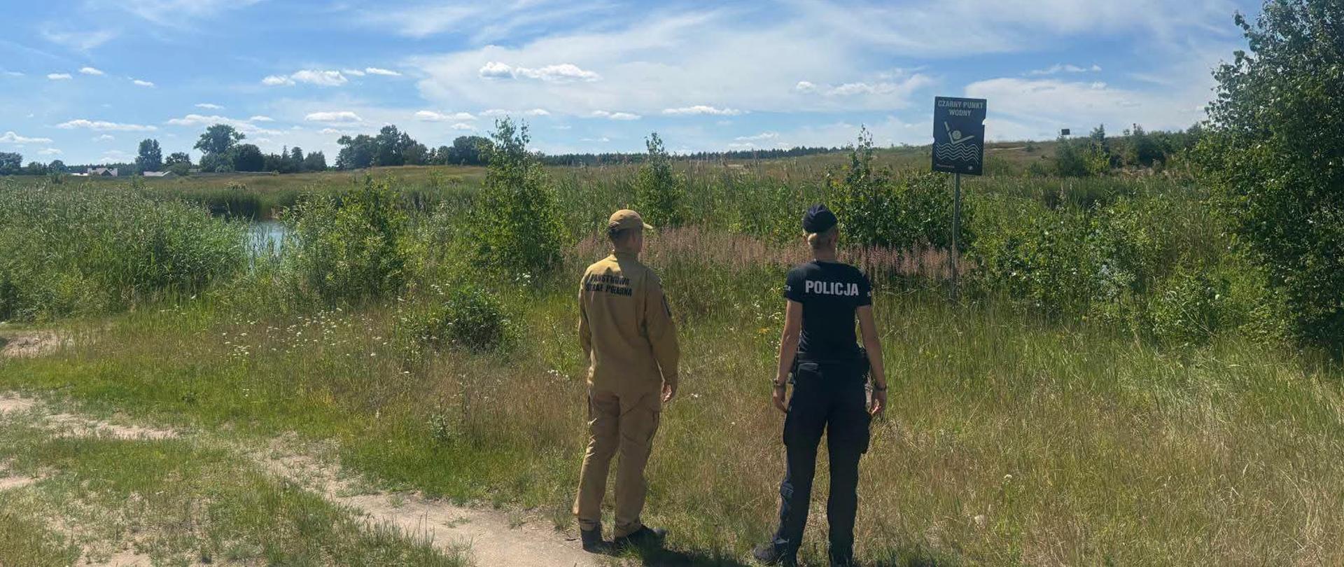 Na zdjęciu widać dwóch umundurowanych mężczyzn – policjanta i strażaka – sfotografowanych od tyłu. Stoją oni na trawiastym, lekko podwyższonym brzegu i patrzą w stronę szerokiego, spokojnego akwenu (jeziora lub stawu).
Po lewej stronie stoi policjant w czarnym, dopasowanym tytule z dużym, białym napisem „POLICJA” na plecach oraz w czarnej czapce z daszkiem.
Po prawej stronie stoi strażak Państwowej Straży Pożarnej w charakterystycznym, piaskowym (beżowym) mundurze z czarnym napisem „PAŃSTWOWA STRAŻ POŻARNA” na plecach oraz w beżowej czapce.
W tle rozciąga się pas zielonych drzew i gęstych szuwarów otaczających wodę. Nad nimi rozpościera się jasnoniebieskie, letnie niebo z delikatnymi, pierzastymi chmurami. W prawym górnym rogu kadru widoczne są liście i gałęzie rosnącego blisko drzewa, które rzuca lekki cień na ziemię. Zdjęcie ma charakter prewencyjnego patrolu służb ratunkowych.