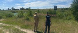 Na zdjęciu widać dwóch umundurowanych mężczyzn – policjanta i strażaka – sfotografowanych od tyłu. Stoją oni na trawiastym, lekko podwyższonym brzegu i patrzą w stronę szerokiego, spokojnego akwenu (jeziora lub stawu).
Po lewej stronie stoi policjant w czarnym, dopasowanym tytule z dużym, białym napisem „POLICJA” na plecach oraz w czarnej czapce z daszkiem.
Po prawej stronie stoi strażak Państwowej Straży Pożarnej w charakterystycznym, piaskowym (beżowym) mundurze z czarnym napisem „PAŃSTWOWA STRAŻ POŻARNA” na plecach oraz w beżowej czapce.
W tle rozciąga się pas zielonych drzew i gęstych szuwarów otaczających wodę. Nad nimi rozpościera się jasnoniebieskie, letnie niebo z delikatnymi, pierzastymi chmurami. W prawym górnym rogu kadru widoczne są liście i gałęzie rosnącego blisko drzewa, które rzuca lekki cień na ziemię. Zdjęcie ma charakter prewencyjnego patrolu służb ratunkowych.
