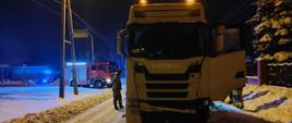 Na pierwszym planie stoi samochód ciężarowy marki SCANIA koloru białego. Ma rozbity przód. W tle stoi strażak oraz samochód gaśniczy na sygnalizacji świetlnej. Aura zimowa w porze nocnej.