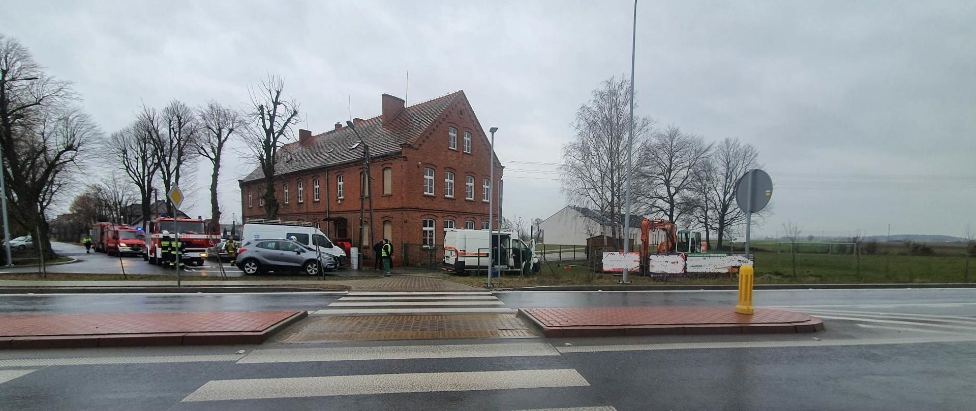Zdjęcie przedstawia pojazdy służb ratowniczych oraz koparkę, która podczas prac ziemnych uszkodziła rurociąg gazowy. W tle budynek Szkoły Podstawowej zbudowany z czerwonej cegły.