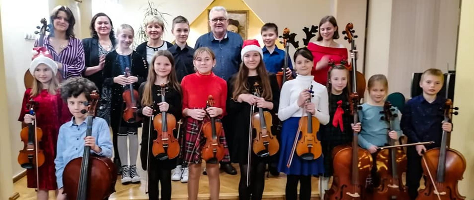 Zdjęcie przedstawia wszystkich wykonawców z koncertu "Koncert smyczkowców" klasy skrzypiec P. Krystyny Ślęzak i wiolonczeli P. Zbigniewa Szuby - 16 grudnia 2022 wraz z nauczycielami P. Krystyną Ślęzak oraz P. Z. Szubą Zdjęcie wykonane w auli PSM I st. w Kłodzku, uczniowie trzymają wiolonczelę lub skrzypce.