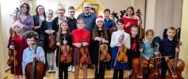 Zdjęcie przedstawia wszystkich wykonawców z koncertu "Koncert smyczkowców" klasy skrzypiec P. Krystyny Ślęzak i wiolonczeli P. Zbigniewa Szuby - 16 grudnia 2022 wraz z nauczycielami P. Krystyną Ślęzak oraz P. Z. Szubą Zdjęcie wykonane w auli PSM I st. w Kłodzku, uczniowie trzymają wiolonczelę lub skrzypce.