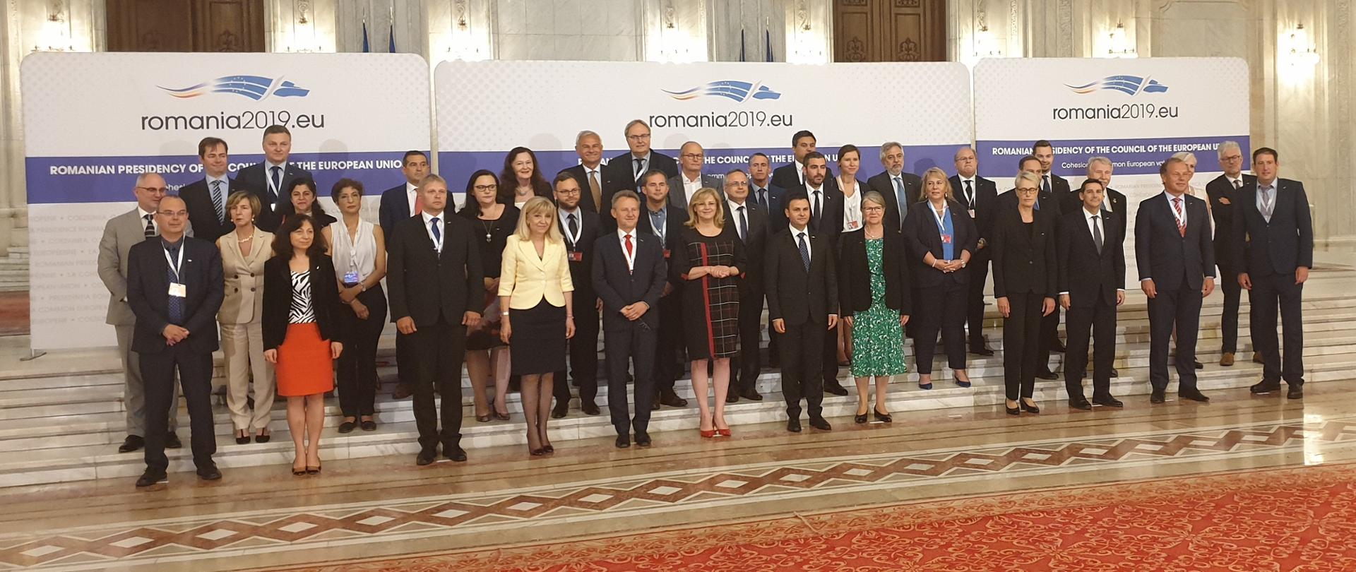 Grupa osób ustawionych w trzy rzędy na schodach, z tyłu ścianki promocyjne z napisem "romania2019.eu".