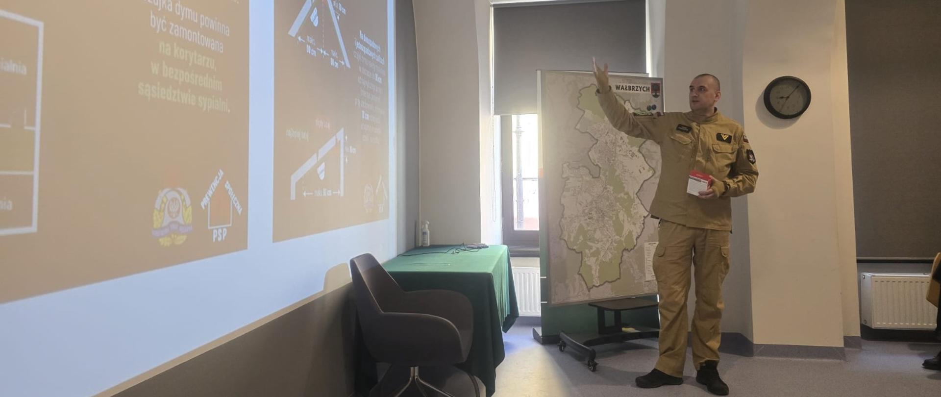 Strażak w piaskowym mundurze służbowym stoi w jasnej sali konferencyjnej. Prawą ręką wskazuje na duży ekran projekcyjny, na którym wyświetlana jest prezentacja w języku polskim pt. „Gdzie umieścić czujkę dymu?”. Slajd zawiera schematy graficzne przedstawiające optymalne miejsca montażu urządzenia na ścianach i sufitach. W tle widoczna jest duża mapa Wałbrzycha na stojaku.