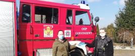 Zdjęcie przedstawia dwie kobiety. Jedna z nich jest strażakiem OSP i wręcza drugiej kobiecie ulotkę dotyczącą szczepień przeciwko COVID-19. W tle kobiet znajduje się czerwony samochód pożarniczy. Widać prawą stronę kabiny pojazdu oraz część zabudowy. Na kabinie znajduje się herb Gminy Rypin, logo ZOSP oraz napis na drzwiach OSP Godziszewy.