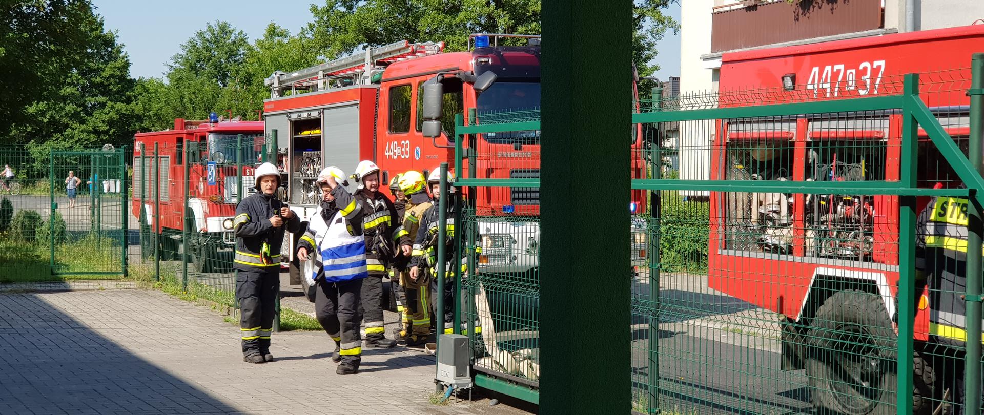Na zdjęciu widać wchodzących na teren przy żłobku strażaków. W tle trzy samochody strażackie ustawione na drodze przy posesji. W tle fragment pobliskiego bloku i przydrożne drzewa. Strażacy w ubraniach specjalnych i hełmach, dowódca w biało niebieskiej kamizelce.
