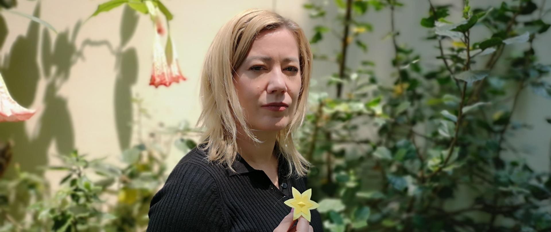 Ambasador Agnieszka Frydrychowicz-Tekieli, ubrana na czarno, patrzy w obiektyw. W tle ogród. W ręku trzyma żółty kwiat – narcyza, symbol powstania w getcie warszawskim.