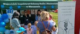 Na zdjęciu widoczny jest namiot Wojewódzkiego Inspektoratu Ochrony Roślin i Nasiennictwa w Warszawie podczas wydarzenia plenerowego. Przed namiotem zgromadzeni są dorośli i dzieci, którzy rozmawiają z pracownicami stoiska. Na stole znajdują się ulotki i materiały informacyjne.