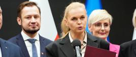 Minister Nowacka stoi i mówi do mikrofonu na stojaku, w ręku trzyma fioletową teczkę.