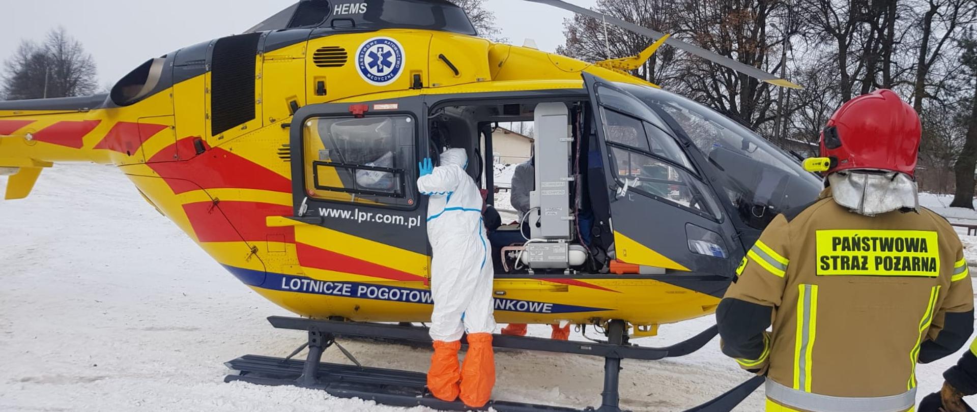 Na zdjęciu widać śmigłowiec LPR. Przy nim ratownik ubrany w ubranie ochronne zabezpieczające przed korona wirusem. Obok stoi strażak.