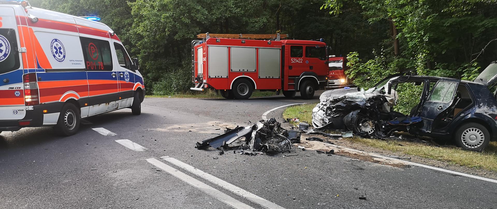 Zdjęcie przedstawia rozbity samochód marki VW Golf znajdujący się na poboczu drogi. W tle samochód pożarniczy oraz karetka pogotowia.