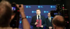 Minister Czarnek stoi obok mężczyzny w garniturze, trzyma w ręku fioletową teczkę, mówi do mikrofonu na stojaku, za nimi niebieska ściana, przed nimi dziennikarze robią zdjęcia.