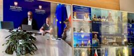 Dwa monitory zawieszone na ścianie. Na jednym widoczny jest podgląd sytuacji w Warmińsko-Mazurskim Urzędzie Wojewódzkim, obejmujący między innymi korytarze urzędu oraz pracowników, w tym zastępcę dyrektora Wydziału Bezpieczeństwa i Zarządzania Kryzysowego oraz inżyniera urzędu. Na drugim monitorze wyświetlony jest podzielony obraz z kilku obszarów monitoringu urzędu.