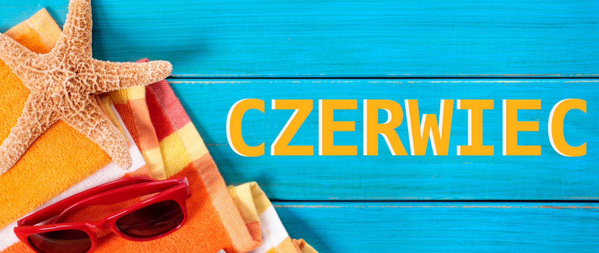 Napis czerwiec na tle niebieskich desek. Obok ręczniki kąpielowe, rozgwiazda i czerwone okulary przeciwsłoneczne. 