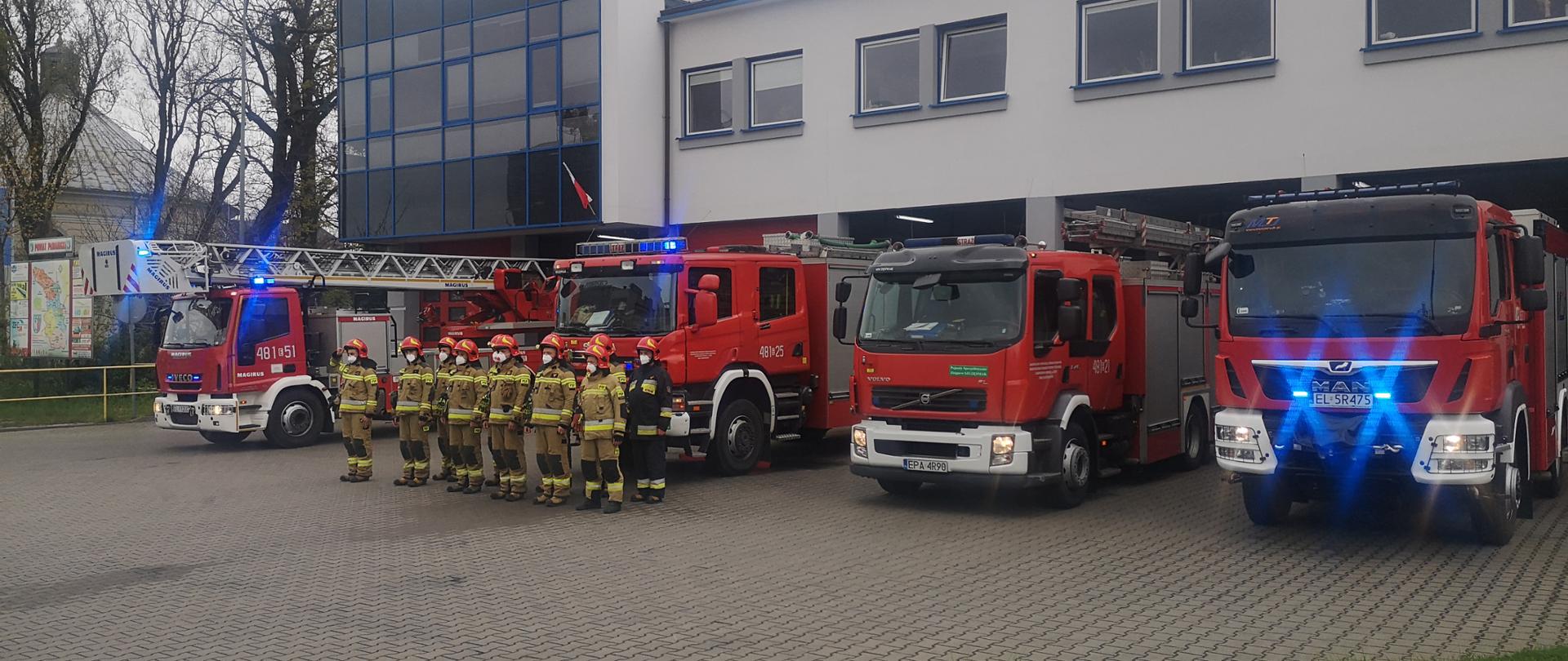 Na zdjęciu widać strażaków w ubraniach bojowych stojących obok samochodów strażackich, samochody stoją przed budynkiem komendy 