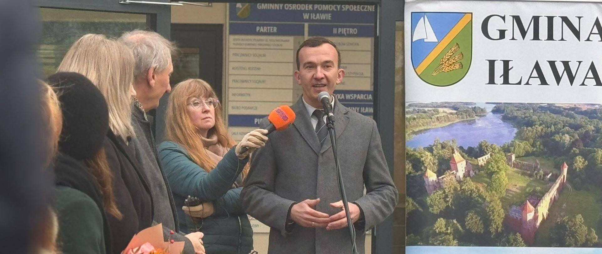 Mężczyzna przemawia na tle nowo otwartego Gminnego Ośrodka Pomocy Społecznej do grupy ludzi stojących na placu 