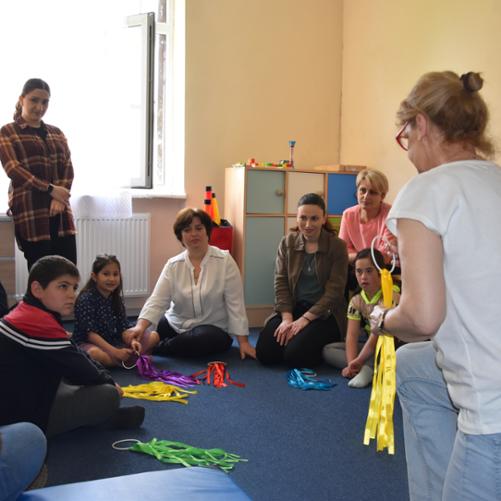 Na podłodze w półkolu siedzą dorośli z dziećmi i patrzą się na instruktorkę, która trzyma w ręku kółko z taśmami; podobne kółka z taśmami leżą na podłodze