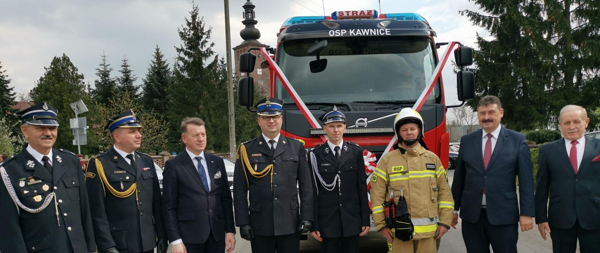grupa strażaków w mundurach wyjściowych stoi obok siebie , za nimi samochód strażacki z biało czerwoną szarfą