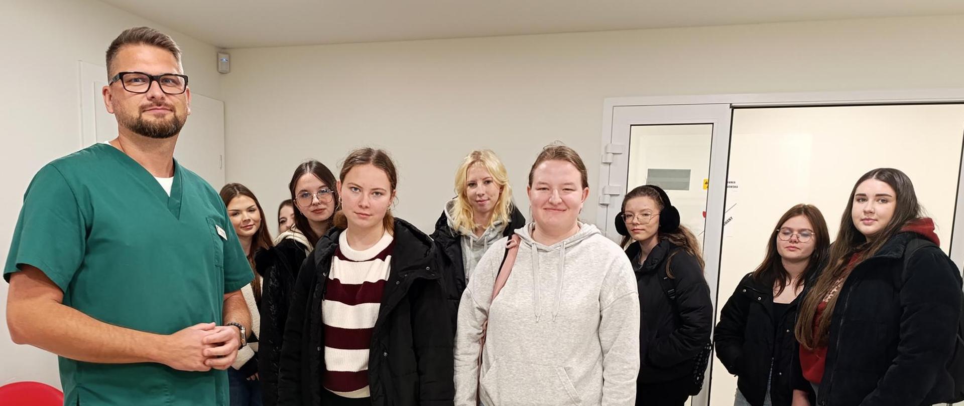 Grupa stojących uczennic, obok mężczyzna w zielonym stroju lekarza.