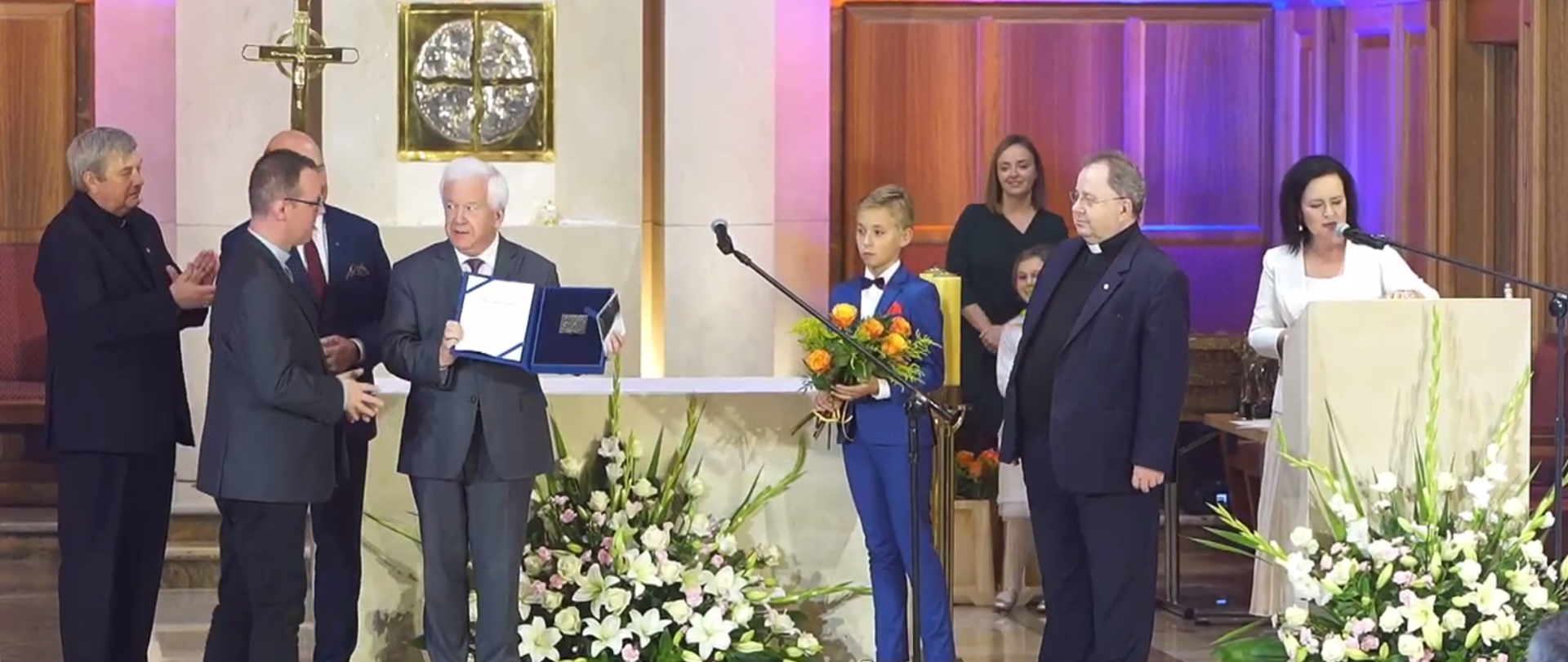 Zdjęcie w poziomie. Grupa osób stojąca na podwyższeniu. W środku prof. Janusz Kawecki prezentujący dyplom.