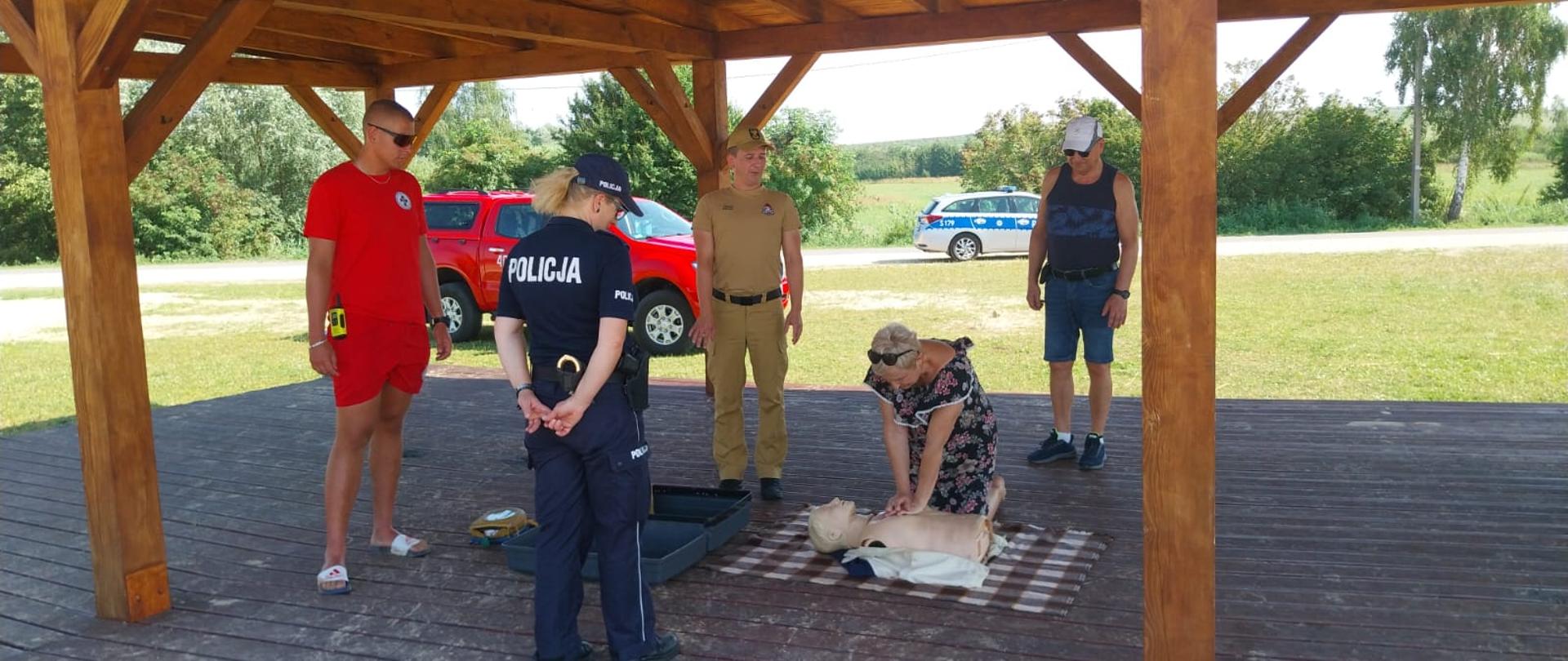 Ukazuje scenę szkoleniową pod drewnianą wiatą na plaży. Kobieta wykonuje resuscytację krążeniowo-oddechową (RKO) na manekinie medycznym. Wokół niej stoi grupa osób przyglądających się ćwiczeniu, w tym ratownik w czerwonej koszulce, funkcjonariuszka policji w mundurze, mężczyzna w mundurze oraz inny obserwator. W tle widać radiowóz policyjny.